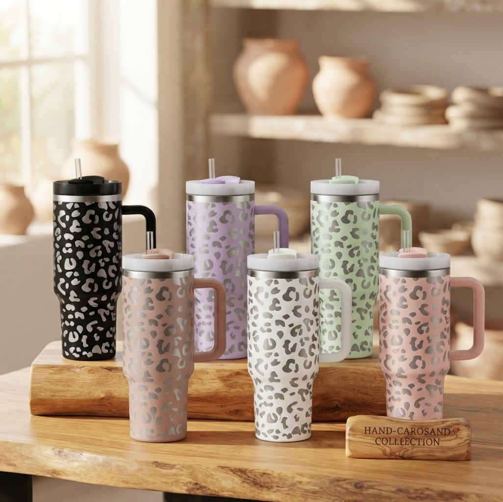 Six insulated tumblers with handles and straws in a soft leopard print pattern, displayed on a wooden table in a warm, rustic shop setting. The tumblers come in black, blush pink, lavender, mint green, and light pink, with gray lids and handles.